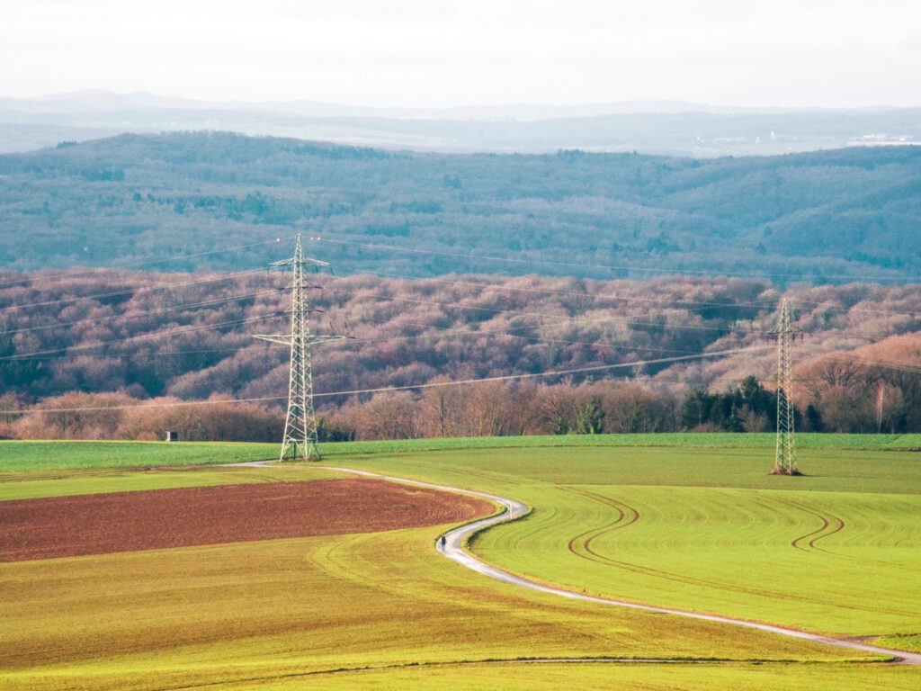 A green field bisected by a meandering dirt road, with power lines and rolling hills in the background.