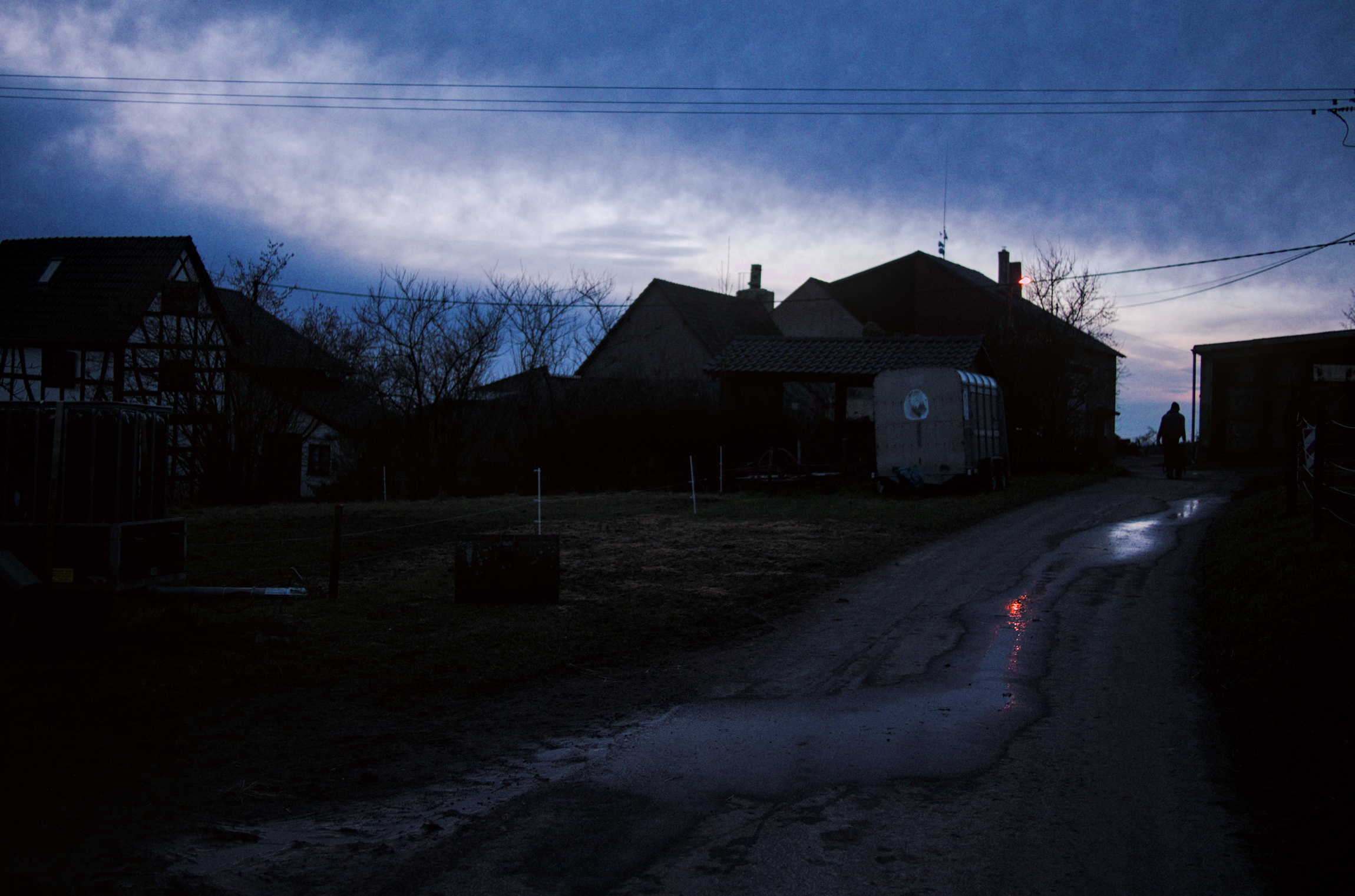 evening scene with a cottage and a barn with a red reflection on a dirt road and a silhouette in the diestance