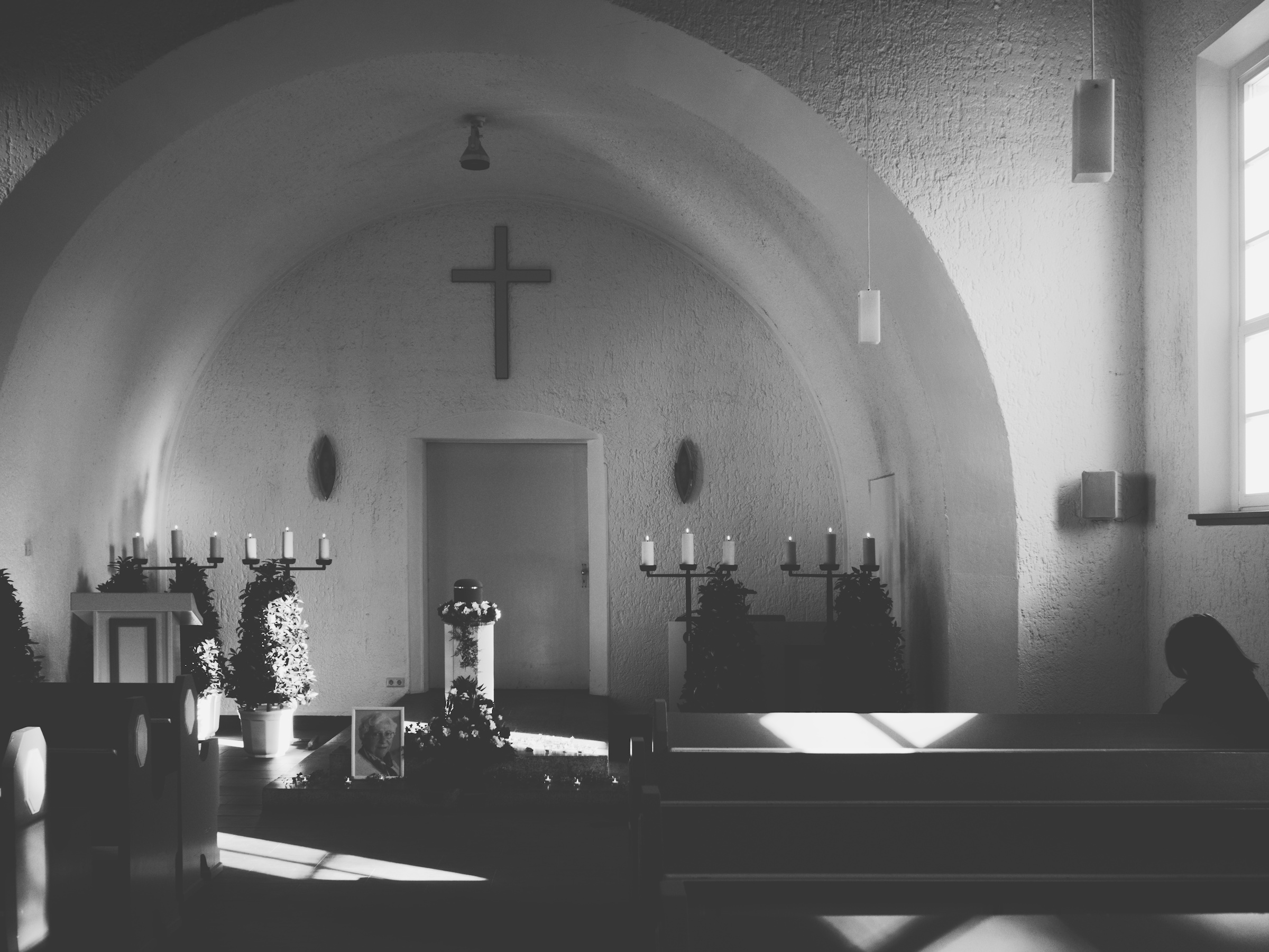 Black and white picture of a chapel interior. At the top end there's an urn, candles and flowers and a portrait on the ground, to the right on the bench there is one single person.