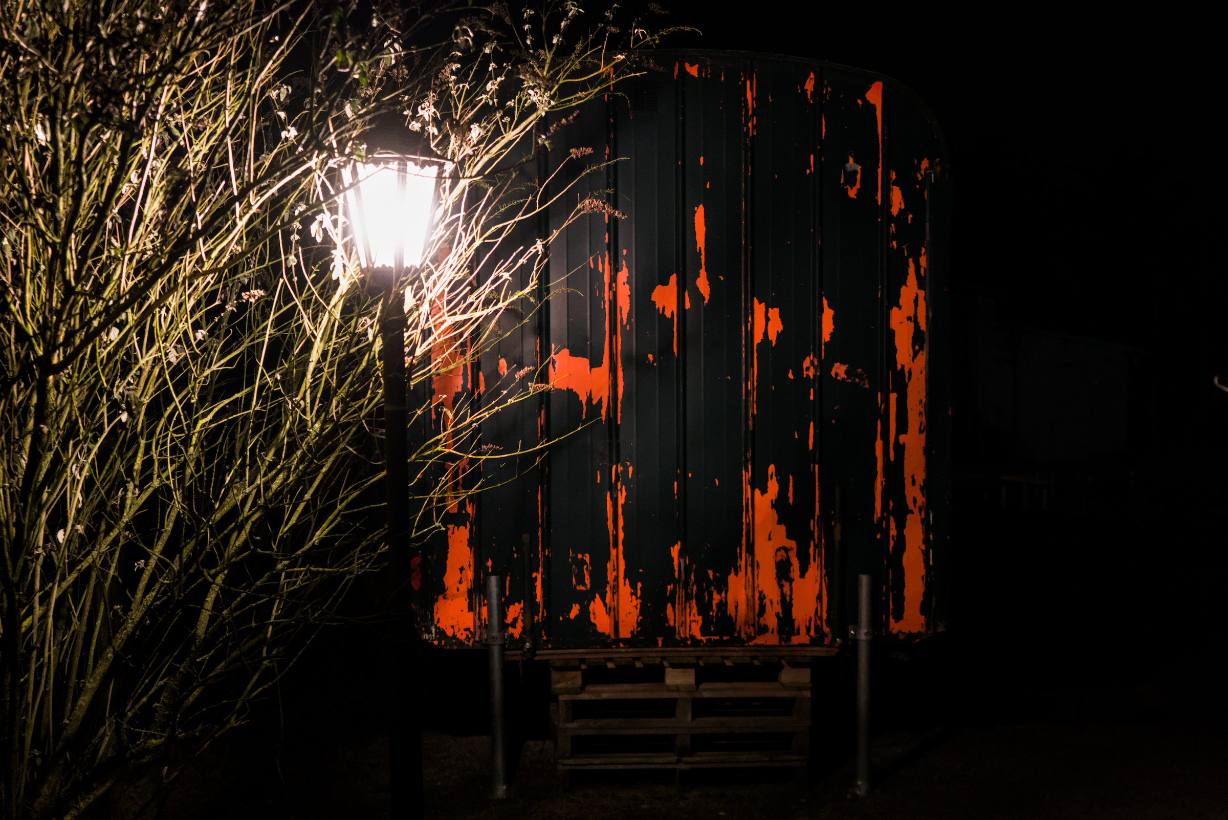 A construction trailer with peeling paint next to a bright streetlamp and a shrub.